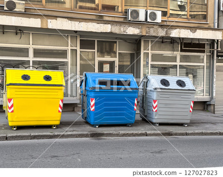 Colorful recycling bins on city sidewalk. Yellow, blue, and gray containers for waste separation in front of building facade. Colorful recycling bins on city sidewalk. Yellow, blue, and gray containers for waste separation in front of building facade. 127027823