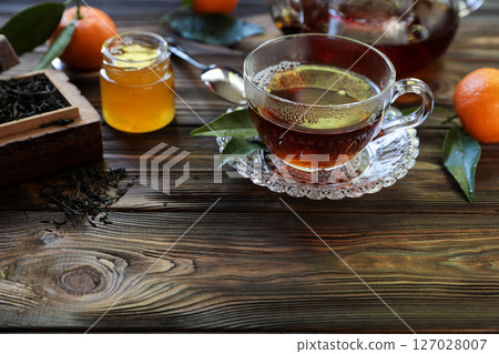Glass cup holds steaming tea accompanied by small jar of honey and fresh oranges. Dried tea leaves spread on wooden surface create a warm atmosphere perfect for afternoon relaxation. Glass cup holds steaming tea accompanied by small jar of honey and fresh oranges. Dried tea leaves spread on wooden surface create a warm atmosphere perfect for afternoon relaxation. 127028007