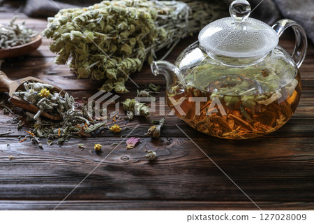 Tea is steeping in glass teapot, surrounded by fresh herbs and dried flowers on weathered wooden table, creating warm and inviting atmosphere for relaxation and enjoyment. Tea is steeping in glass teapot, surrounded by fresh herbs and dried flowers on weathered wooden table, creating warm and inviting atmosphere for relaxation and enjoyment. 127028009