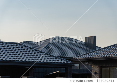 Metallic tiles cover the rooftops of several modern houses, reflecting sunlight under a clear blue sky in a calm suburban neighborhood 127029253