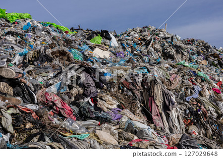 A vast accumulation of discarded clothing, plastic, and debris fills the landfill, highlighting urgent issues of pollution and waste management 127029648