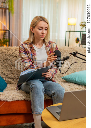 Woman preparing to record podcast on laptop, adjusting microphone checking notes with nervous focus 127030031