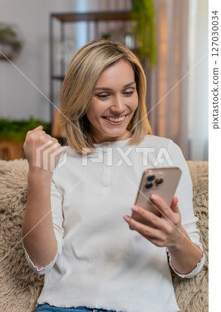 Adult smiling woman texting happily on smartphone at home after winning online lottery jackpot 127030034