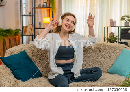 Adult woman dancing on home sofa with wireless headphones, enjoying favorite song energetic mood 127030038