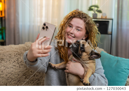 Young woman taking selfie with pug dog on home sofa, smiling with joy while holding cute pet indoors 127030043