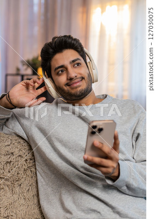 Young Indian man guy on sofa wearing wireless headphones listening to music on smartphone at home 127030075