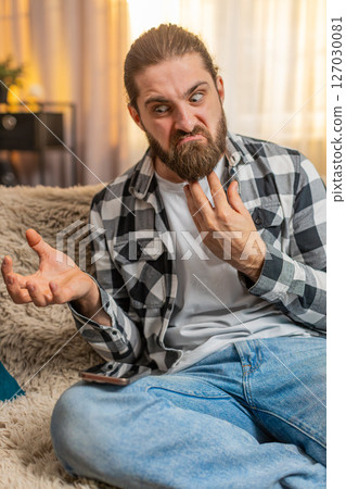 Young man anxious at home, urgent phone call on knee, frustration, loud speaker, intense quarrel 127030081