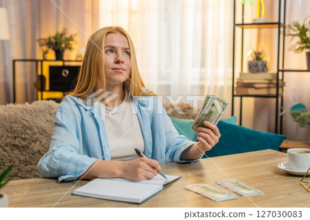 Young woman organizing expenses at home, counting money cash banknotes, writing notes, happy focus 127030083