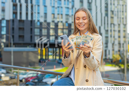 Happy rich mature Caucasian businesswoman using smartphone, holding euro money cash on city street Happy rich mature Caucasian businesswoman using smartphone, holding euro money cash on city street 127030102