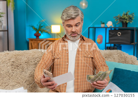 Mature man on sofa using smartphone calculator, counting cash and bills, feeling exhausted at home Mature man on sofa using smartphone calculator, counting cash and bills, feeling exhausted at home 127030155