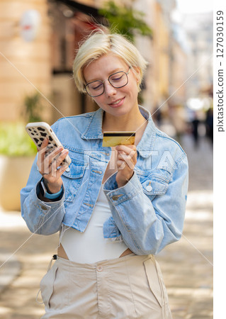 Happy Caucasian woman using credit card smartphone while purchases online shopping on city street Happy Caucasian woman using credit card smartphone while purchases online shopping on city street 127030159