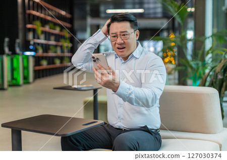 Upset businessman using smartphone becomes shocked sudden lottery results in modern office lobby 127030374