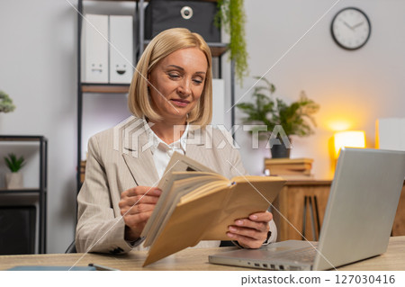 Mature businesswoman reading interesting book smiling enjoying literature taking a rest after work Mature businesswoman reading interesting book smiling enjoying literature taking a rest after work 127030416