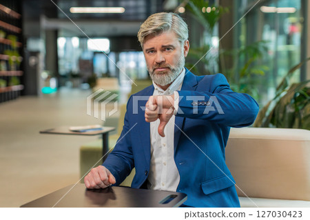 Upset Caucasian businessman in suit showing thumb down sign gesture sitting in modern office lobby 127030423