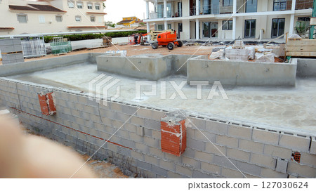 A new swimming pool is taking shape near an unfinished house, with construction materials in the background and workers present on the site. The setting is a suburban neighborhood. A new swimming pool is taking shape near an unfinished house, with construction materials in the background and workers present on the site. The setting is a suburban neighborhood. 127030624