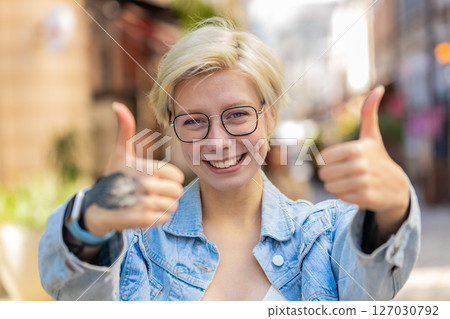 Cheerful young European woman showing double thumbs up gives positive reply standing on city street Cheerful young European woman showing double thumbs up gives positive reply standing on city street 127030792