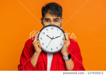 Serious strict Indian man holding office clock hiding behind, checking time on watch, obscuring face 127030812