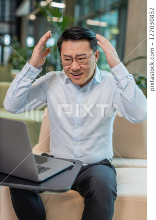 Sad businessman using laptop loses becoming shocked sudden lottery results in modern hotel lobby Sad businessman using laptop loses becoming shocked sudden lottery results in modern hotel lobby 127030832