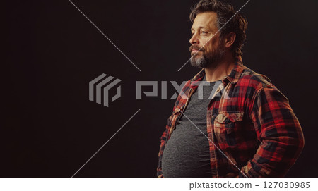 pregnant man in plaid shirt stands with hands on hips against dark background. Concept of confidence and casual style. 127030985
