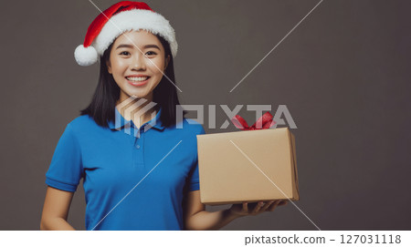 Smiling woman in a Santa hat holds a gift box with a red ribbon. Concept of festive celebration and holiday spirit. 127031118