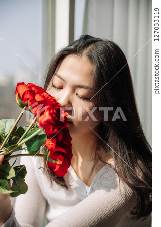 Asian girl smelling red roses by the window Asian girl smelling red roses by the window 127033119