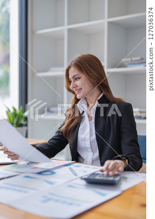 Attractive woman reviews financial documents and calculates figures at desk. 127036374