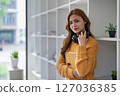 Young pretty woman leans on bookshelf holding a book and headphones. 127036385