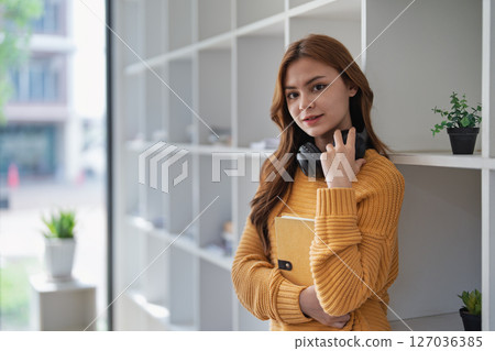 Young pretty woman leans on bookshelf holding a book and headphones. Young pretty woman leans on bookshelf holding a book and headphones. 127036385