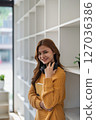 Smiling young woman holds a book in front of a white bookshelf indoors. 127036386