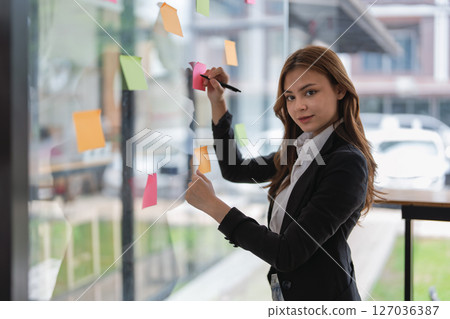 Businesswoman writing ideas on sticky notes on glass wall in office space. 127036387