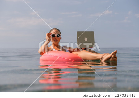 A woman is floating on a pink inflatable raft in the ocean while holding a laptop. Concept of relaxation and leisure, as the woman is enjoying her time in the water while working on her laptop. A woman is floating on a pink inflatable raft in the ocean while holding a laptop. Concept of relaxation and leisure, as the woman is enjoying her time in the water while working on her laptop. 127038415