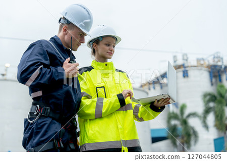 Man and woman petrochemical or gas factory workers or technicians work using laptop with man talk using walkie talkie and they stand in front of tank in factory. 127044805