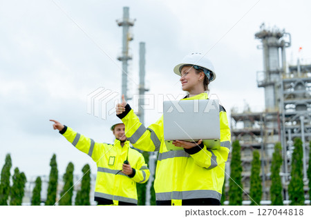 Petrochemical factory worker point to his right side with one coworker woman use laptop to work in front and they stay with factory in the background. Petrochemical factory worker point to his right side with one coworker woman use laptop to work in front and they stay with factory in the background. 127044818