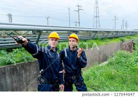 Two petrochemical or gas factory workers discuss about work with one point to their right side and stand in front of pipeline of factory. 127044847