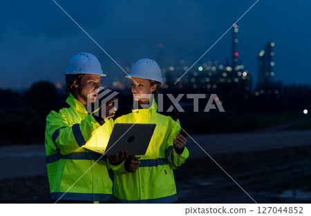 Two petrochemical workers or engineers discuss about work using laptop at night and stand in front of the factory with light. 127044852