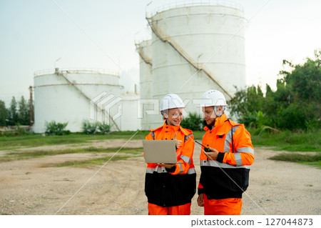 Wide shot of technician or gas factory worker woman hold and use laptop to work with coworker man in front of gas or petrochemical tank and they look happiness with their work. 127044873