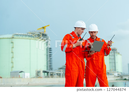 Two engineers or gas factory technicians hold walkie talkie and laptop to discuss about their work and stand in front of tank near the bay with day light. 127044874