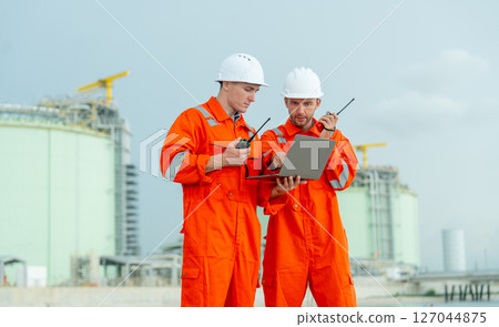 Close up two engineers or gas factory technicians hold walkie talkie and laptop to discuss about their work and stand in front of tank near the bay with day light. 127044875
