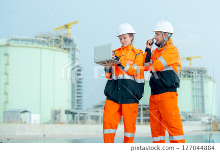 Lower view of two petrochemical or gas factory workers man and woman stand in front of factory tank near the bay and work together using laptop. Lower view of two petrochemical or gas factory workers man and woman stand in front of factory tank near the bay and work together using laptop. 127044884