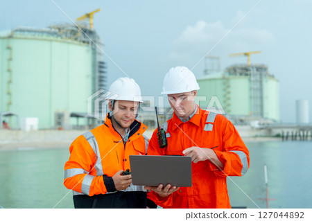 Two petrochemical or gas factory workers men stand in front of factory tank near the bay and work together using laptop. 127044892