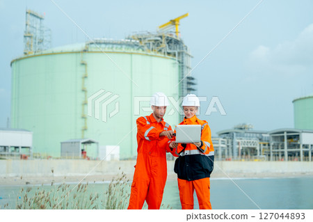 Two petrochemical or gas factory workers man and woman or engineers stand in front of factory tank near the bay and work together using laptop. Two petrochemical or gas factory workers man and woman or engineers stand in front of factory tank near the bay and work together using laptop. 127044893