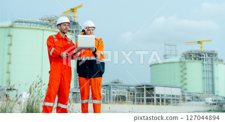 Wide shot of two petrochemical or gas factory workers man and woman or engineers stand in front of factory tank near the bay and work together using laptop. 127044894