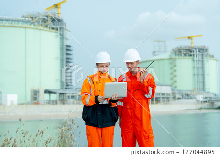 Two petrochemical or gas factory workers man and woman stand in front of factory tank near the bay and work together using laptop. 127044895