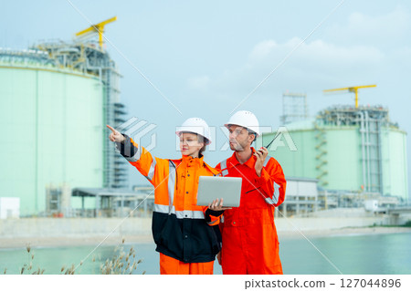 Petrochemical or gas factory workers man and woman work together with one hold laptop and point to ride side and man hold walkie talkie stay in front of factory tank near the bay. Petrochemical or gas factory workers man and woman work together with one hold laptop and point to ride side and man hold walkie talkie stay in front of factory tank near the bay. 127044896