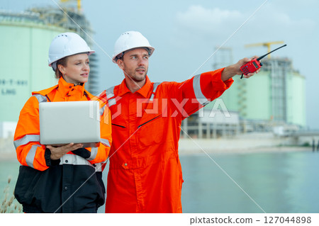 Petrochemical or gas factory workers man and woman work together with one hold laptop and look at direction of man point to left side in front of factory tank near the bay. 127044898