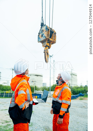 Vertical image of two factory workers or technician man and woman look at part of crane in front of tank in factory workplace. 127044914