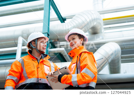 Two petrochemical or gas factory workers man and woman discuss about work using laptop and stand near pipeline in factory workplace. Two petrochemical or gas factory workers man and woman discuss about work using laptop and stand near pipeline in factory workplace. 127044925