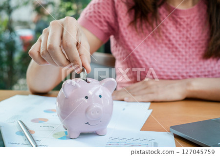 Close-up of a woman placing a coin into a pink piggy bank, surrounded by financial documents and a laptop on a wooden desk. 127045759