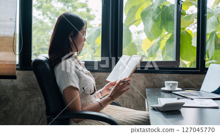 A woman sits by a window reading a book in a modern office, surrounded by greenery and natural light. A woman sits by a window reading a book in a modern office, surrounded by greenery and natural light. 127045770