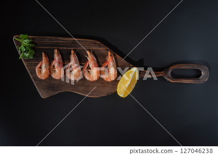 Boiled shrimps on a cutting board. Serving shrimps with lemon and herbs. Red shrimps on a black background. 127046238